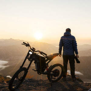 Rider standing beside a Freego electric dirt bike on a mountain overlook at sunrise, showcasing off-road adventure and scenic outdoor exploration.