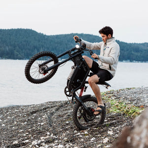 Rider performing a wheelie on a Freego electric dirt bike along a rocky lakeshore, demonstrating powerful off-road performance and control.