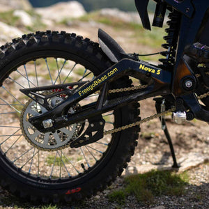 Close-up of the rear wheel and chainstay of a black freego nova 5 dirt bike on a rocky outdoor trail. The frame shows 'FreecoBikes.