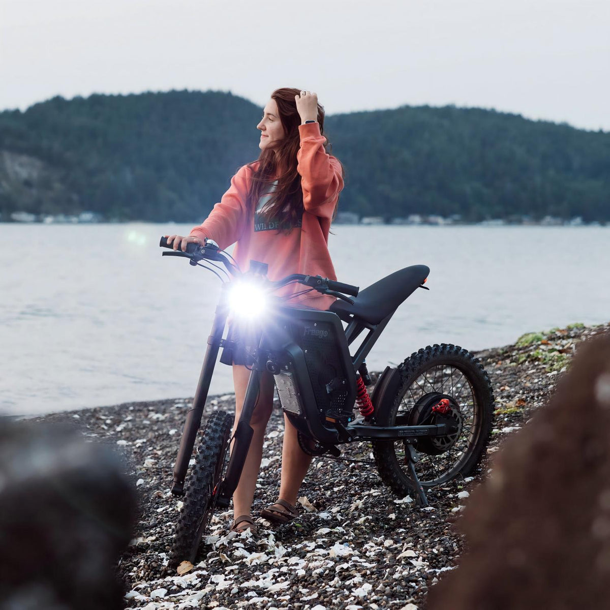 Woman standing beside a Freego electric dirt bike on a rocky lakeshore, enjoying a peaceful outdoor adventure with scenic waterfront views.