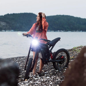 Woman standing beside a Freego electric dirt bike on a rocky lakeshore, enjoying a peaceful outdoor adventure with scenic waterfront views.