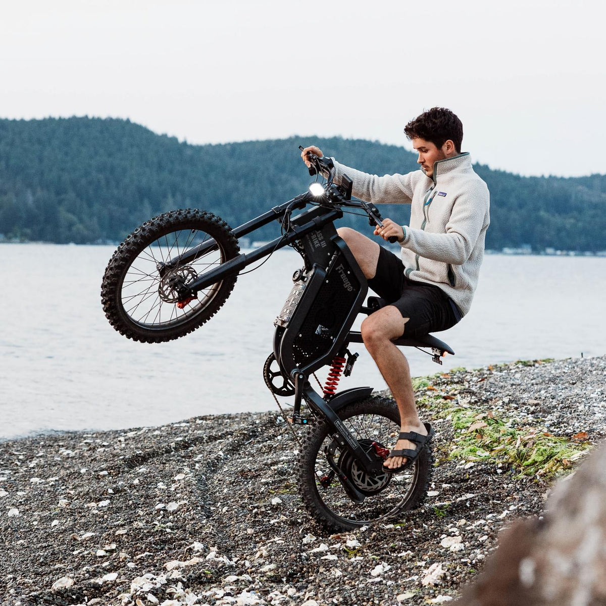 Rider performing a wheelie on a Freego electric dirt bike along a rocky lakeshore, demonstrating powerful off-road performance and control.