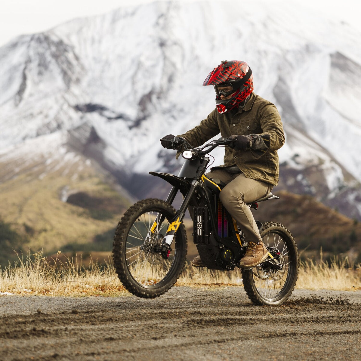 Dynamic outdoor riding shot of rider with red helmet lifting front wheel on Freego N1 electric dirt bike, snow-capped mountain range backdrop on dirt trail