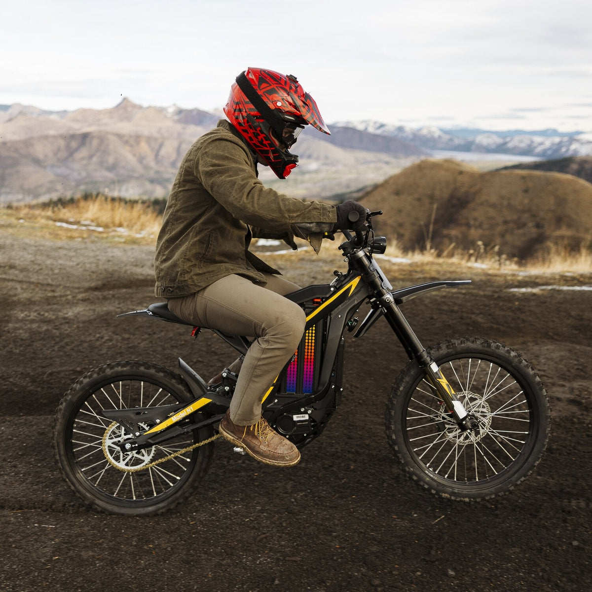 Side profile outdoor riding shot of rider with red helmet on Freego N1 electric dirt bike riding over mountain dirt terrain, rolling hills landscape background