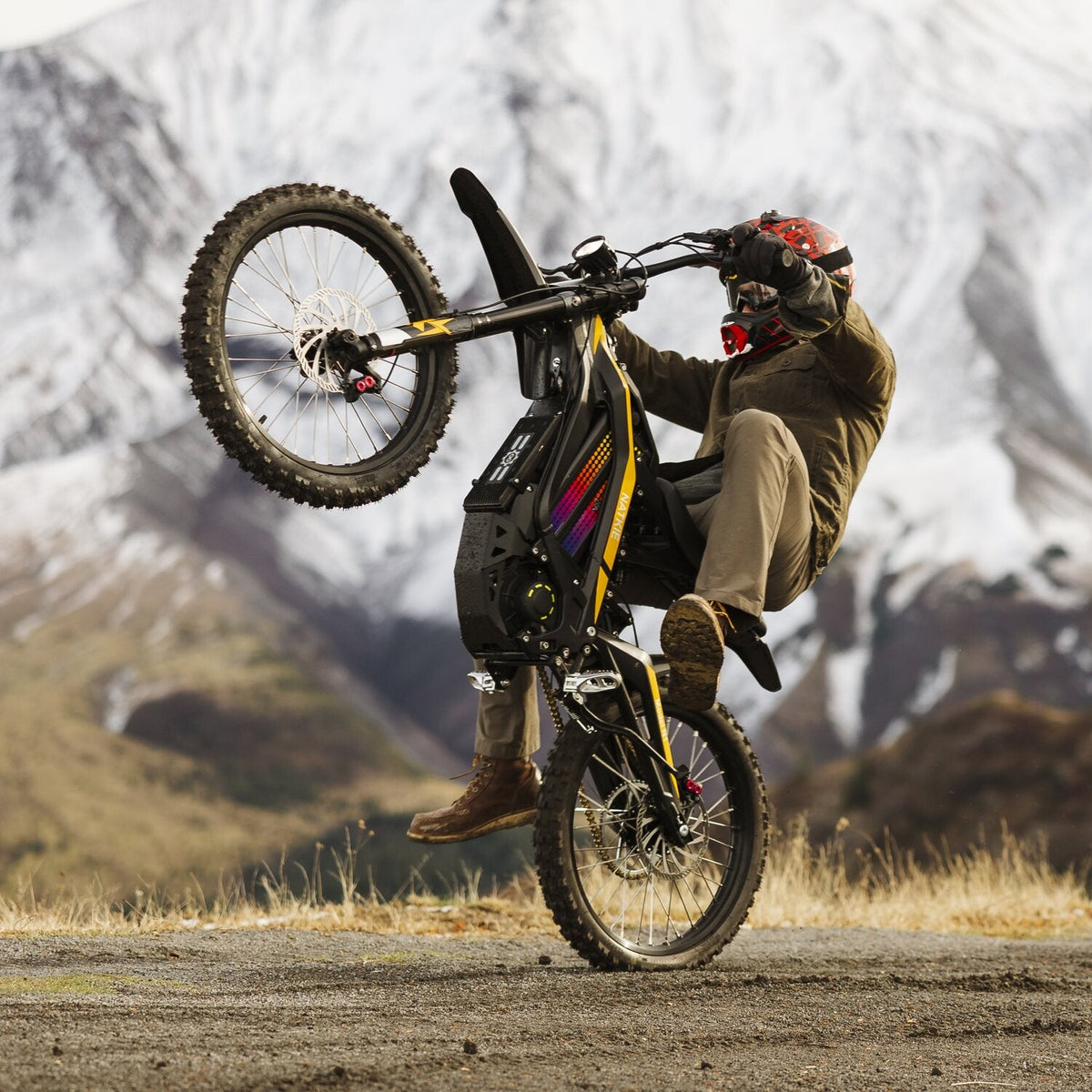 Dynamic stunt action shot of rider performing wheelie on Freego N1 electric dirt bike, rugged mountain dirt trail with snow-capped mountain backdrop