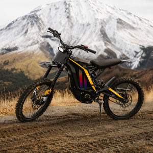 Static product shot of Freego N1 electric dirt bike with yellow graphic decals, parked on mountain dirt road with snow-capped mountain range scenery background