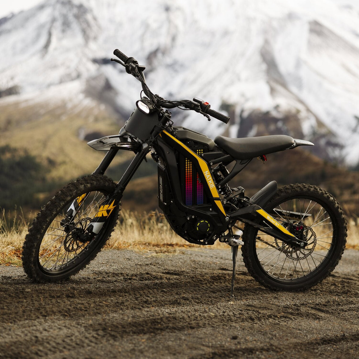 Static outdoor product shot of Freego N1 electric dirt bike with yellow decorative stripes, parked on mountain dirt ground with snow mountain background