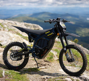 A black and blue freego nova 5 electric dirt bike with yellow accents is parked on a rocky mountain ridge with a scenic mountain valley in the background.