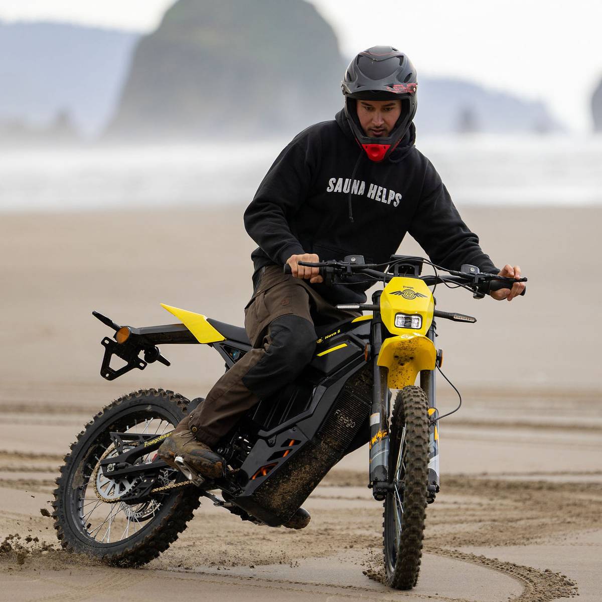A person wearing a black helmet and black hoodie rides a yellow and black feego nova 5 electric dirt bike on a sandy beach with ocean stacks in the background.
