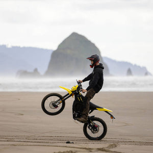 A person riding a yellow and black freego nova 5 dirt bike on a sandy beach with ocean waves and large rock formations in the background, performing a wheelie.