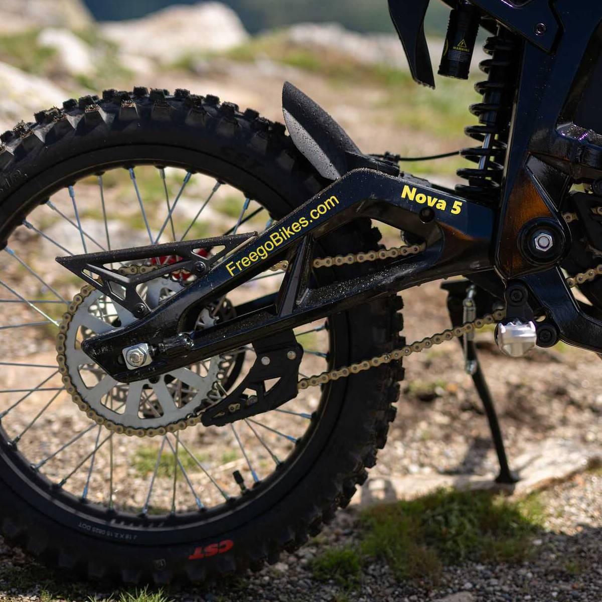 Close-up of the rear wheel and chainstay of a black freego nova 5 dirt bike on a rocky outdoor trail. The frame shows 'FreecoBikes.