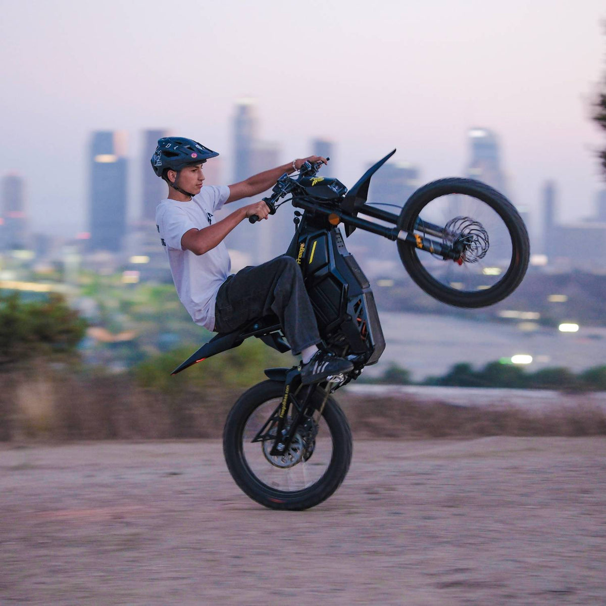 Rider performing wheelie on Freego Nova5 electric dirt bike with city skyline background at dusk
